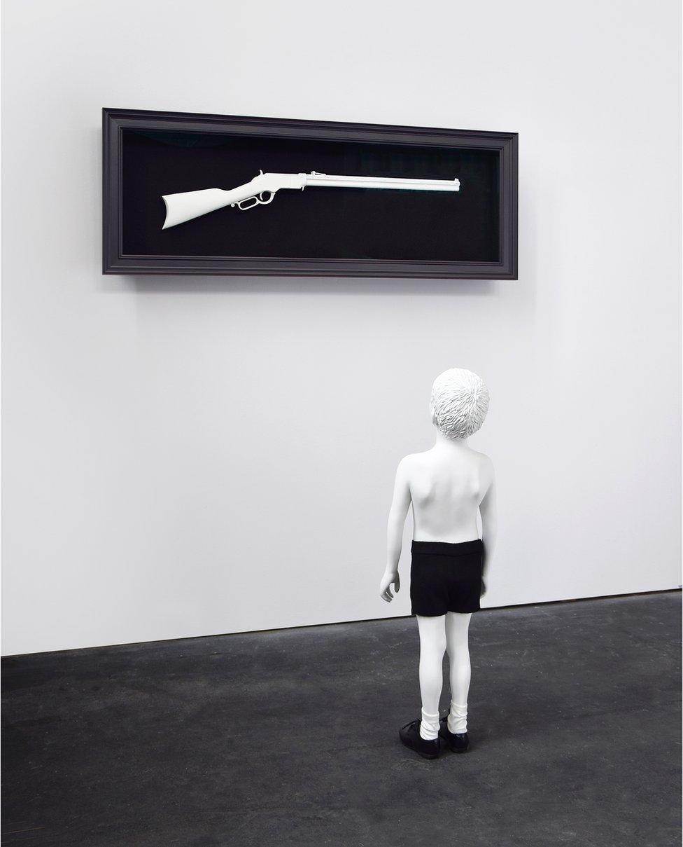 One Day (2015) is a monchrome sculpture in which a boy looks up at a rifle mounted on the wall