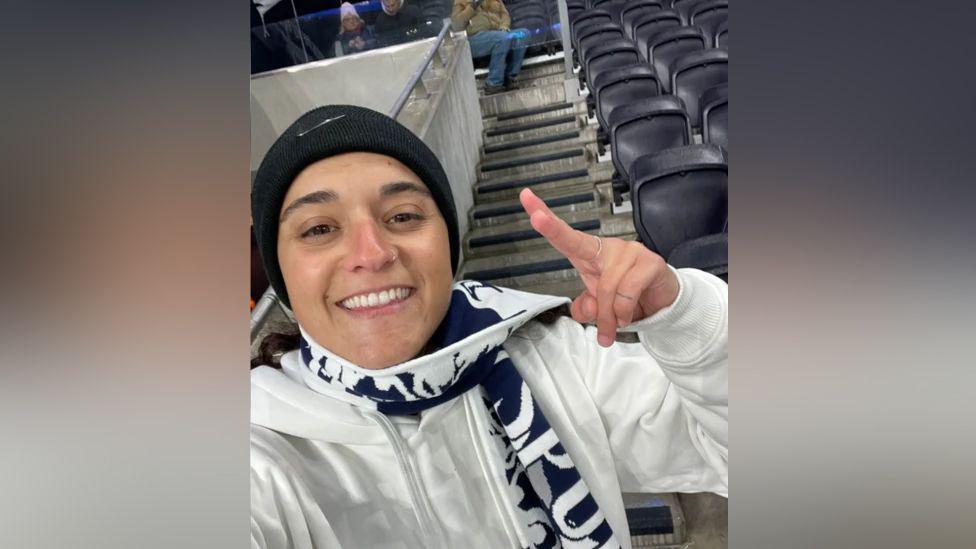 Emily Damari, a young woman wearing a Spurs scarf and a black beanie hat, smiles in the stands of a football ground