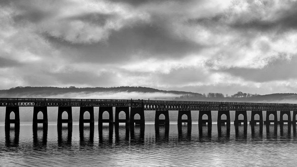 A black and white photo showing the arches holding up the Tay Rail Bridge. There is water underneath. There is a cloudy sky.