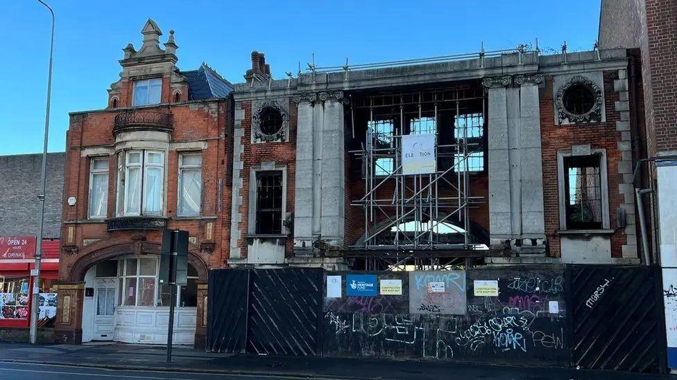 A ruined early 20th Century cinema, built in brick and stone, with blackened windows and scaffolding across the front.