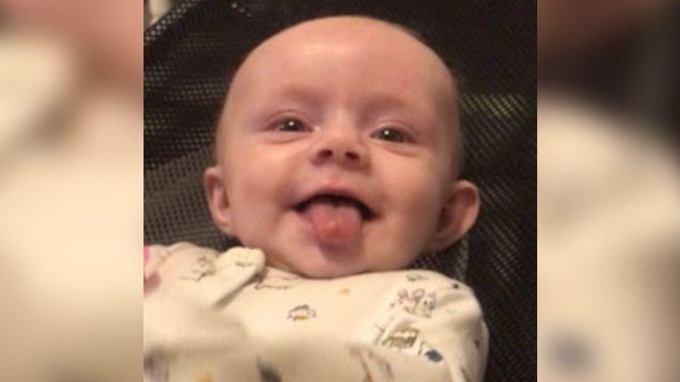 Lexi wearing a white baby grow with tiny mice on it. She is lying down and looking up towards the camera with her tongue out in a cheeky smile.