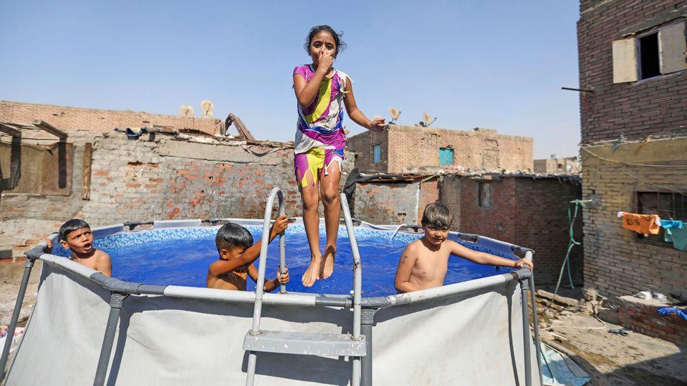 Three children are in a paddling pool and one child is outside it. They are outside and there are some dilapidated buildings in the background in Cairo, Egypt - Tuesday 26 August 2025.
