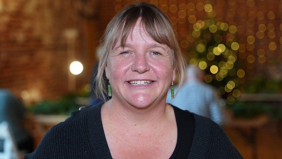 Heather Deane who has fair hair cut in a  fringe and pulled back behind her head, with some strands falling around her ears. She is wearing a dark v-necked top under a black cardigan