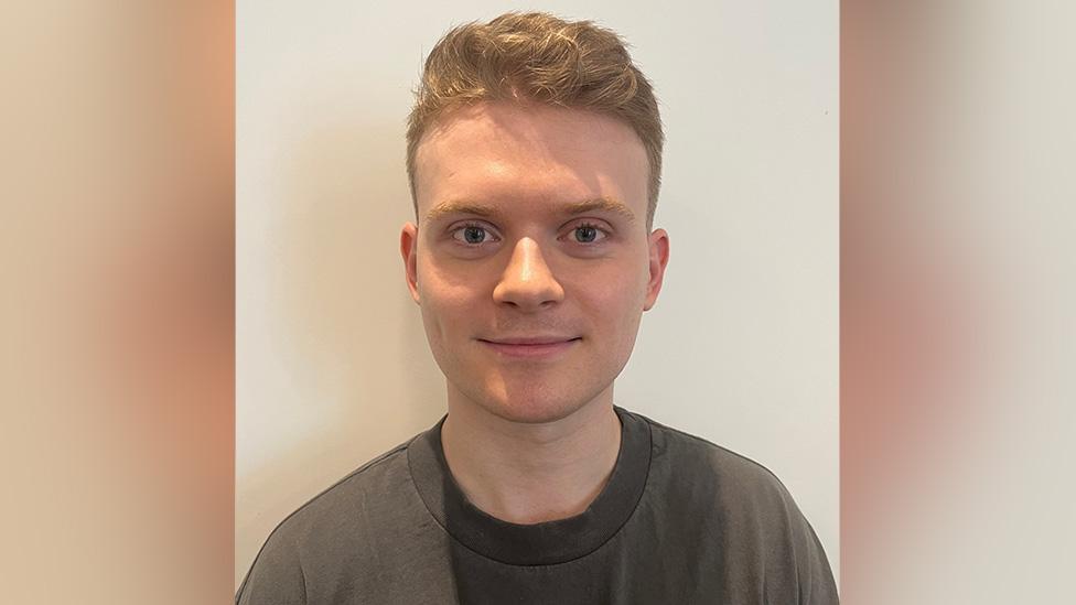 A young white man stands in front of a white wall wearing an olive green sweatshirt. He's got short, orangey-blonde hair that's shaved at the sides but longer on top. He's got a smooth complexion with a dimple on his right cheek, and large blue eyes. He's smiling ever so slightly, facing the camera straight-on as if posing for a passport photograph.