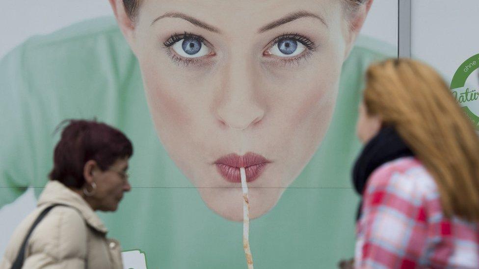 Women walk in front of a commercial featuring a woman eating pasta, outside a restaurant in Berlin