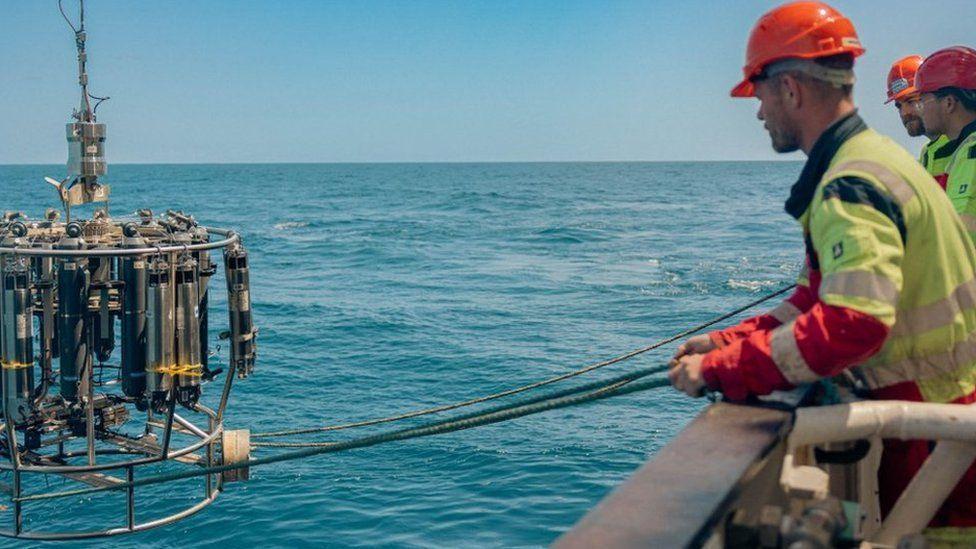 Three men in high vis jackets, wearing hard hats are pulling in the instruments, that measure the AMOC, from the ocean using ropes.