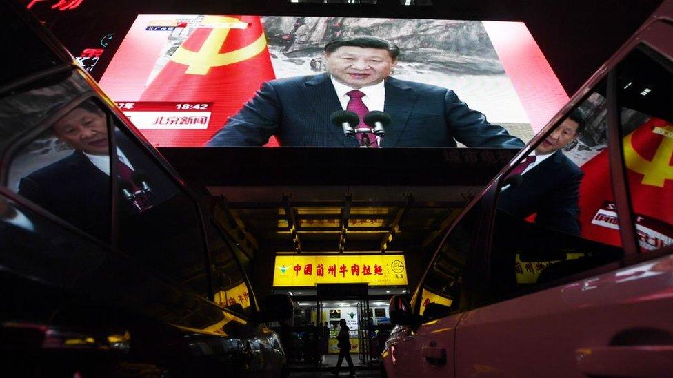 People walk outside a shop below a screen showing news coverage about Chinese President Xi Jinping.