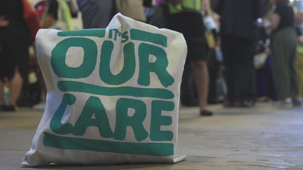 A bag with It's Our Care written on it on the floor with people behind