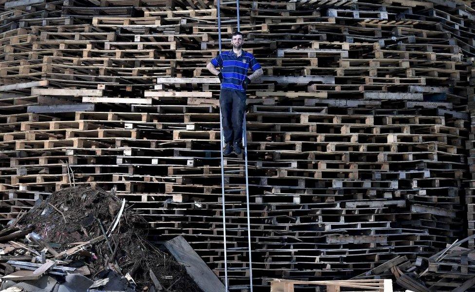 'Hurka' the chief architect and builder of the Craigyhill bonfire takes a break from stacking pallets to pose with his creation whilst balancing on a ladder on July 10, 2017 in Larne,