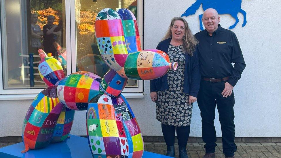 Rainbow coloured dog statue with two people stood to the right of it