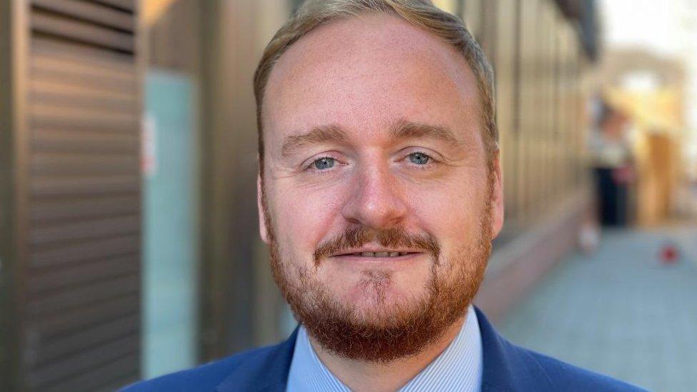 Man with short ginger hair and beard wearing a suit outside a council building