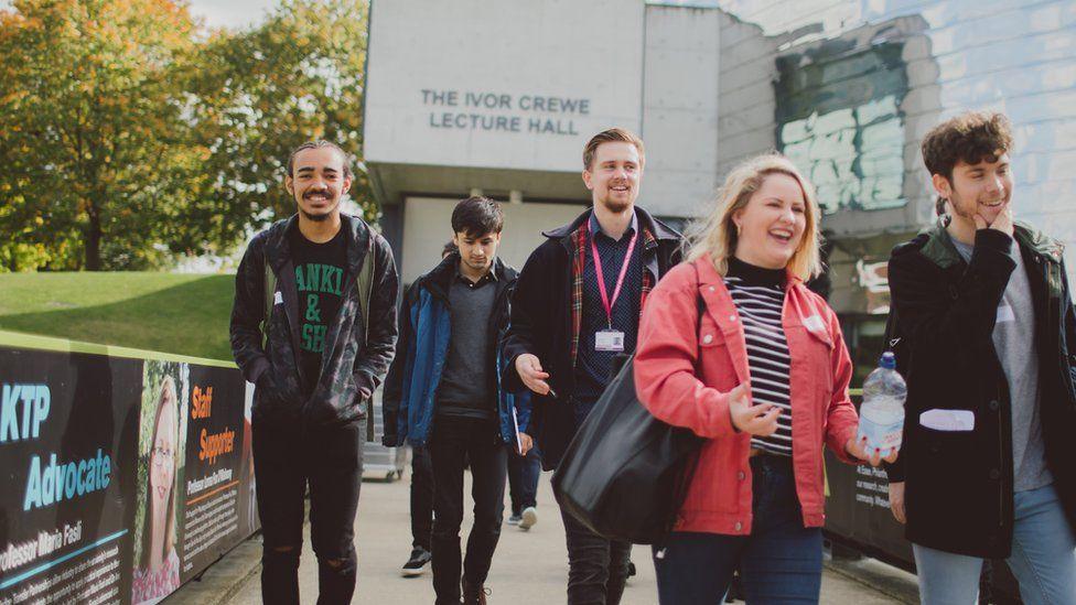 Five students at the University of Essex walk outside a lecture hall. The building is silver and has trees next to it. The students are all wearing coats and smiling