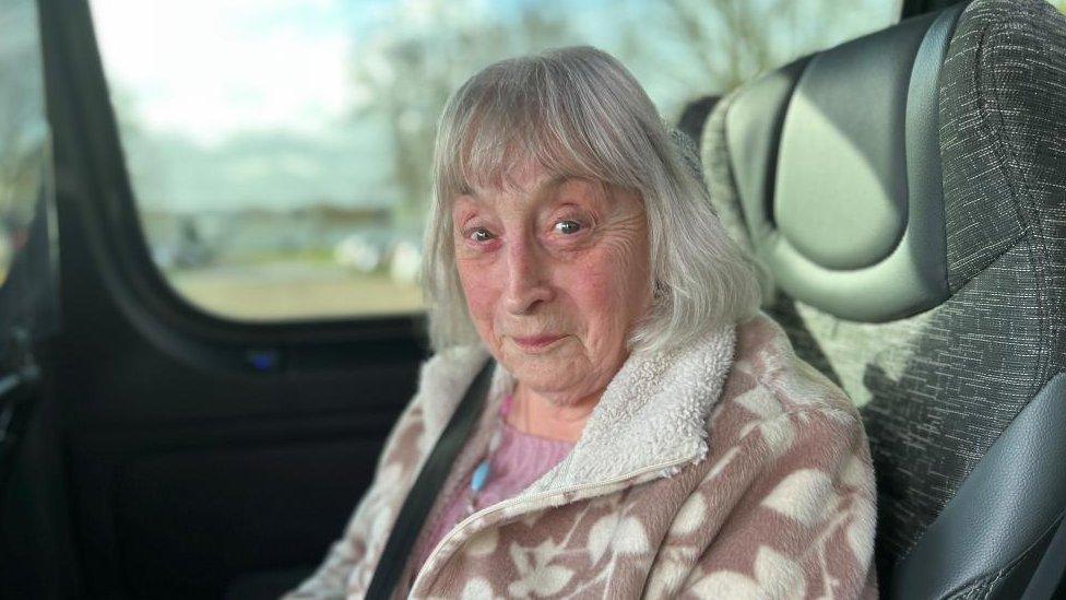 Woman with grey hair wearing a brown and white coat sitting on a bus