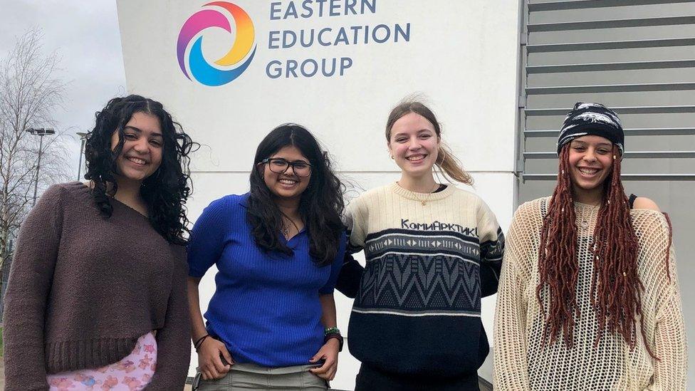 Four teenage girls stand in front of a school sign