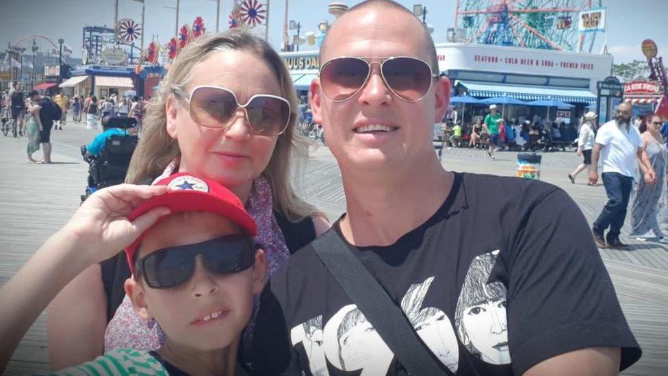 A man and woman with a young boy, all wearing sunglasses, posing for the camera while at a boardwalk fairground.