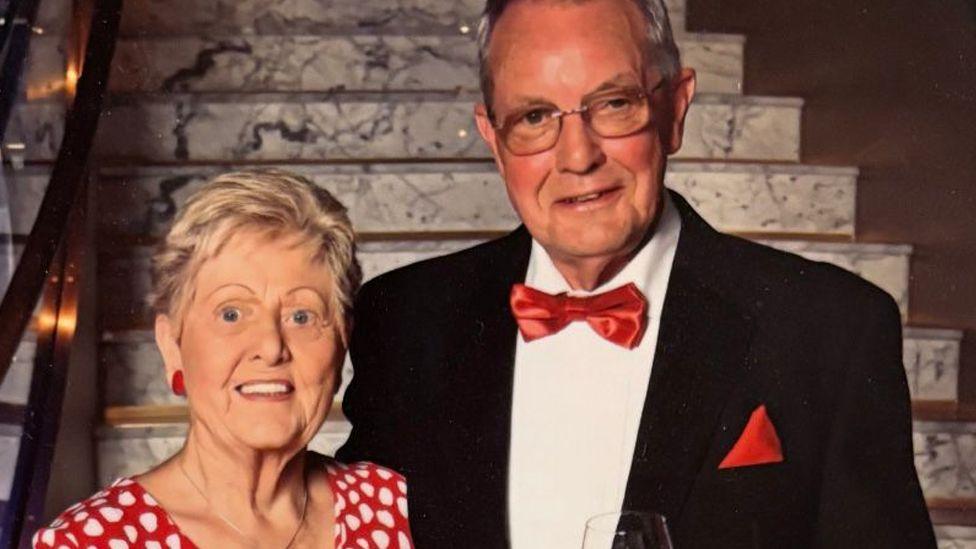 Close-up picture of a smiling Marilyn and Ron Hill in formal dress in front of marble steps. Marilyn is wearing a red dress with white spots and Ron is wearing a black suit with a red dickie bow and red handkerchief in his top pocket.
