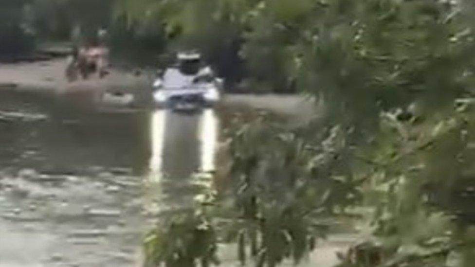 A second car up to its wheels in water driving along the lakeside