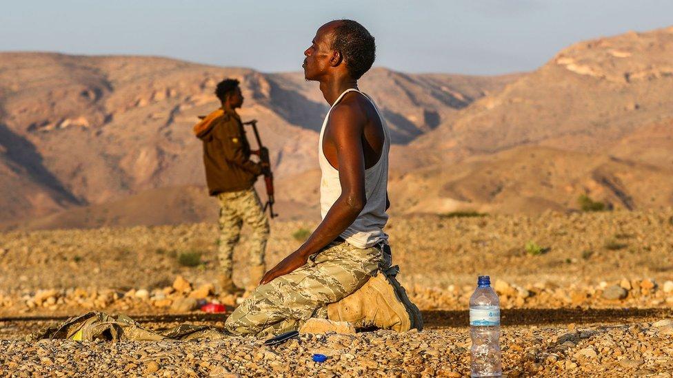 A man stands guard on the left and another prays in the middle of the mountains