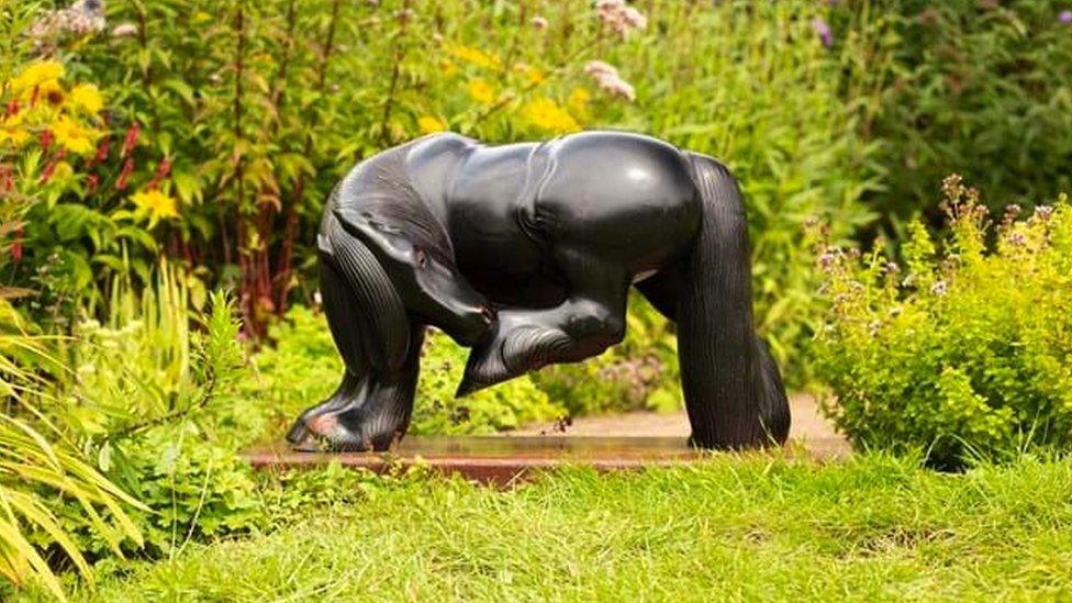 Image of the Black Mare sculpture. It is a mahogany horse, turning to nip at a fly on its rear. The sculpture has been photographed amongst grass and flowers.