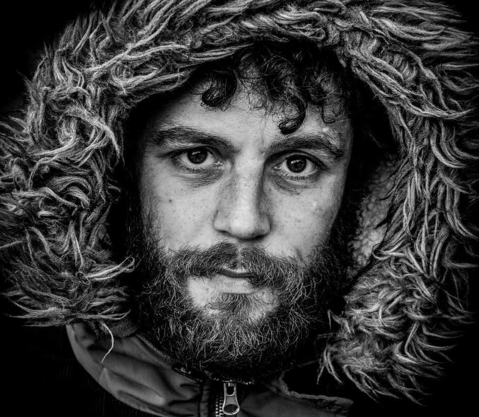 Black and white image of bearded man with fur lined hood up