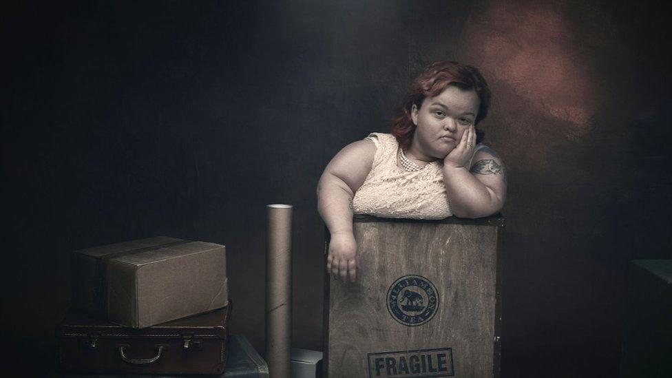 A woman with dwarfism has a bored expression on her face as she stands in a box marked fragile