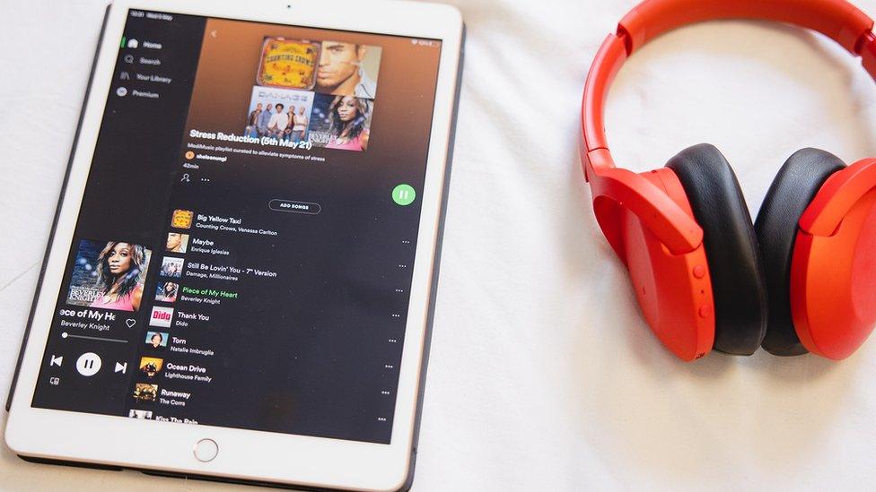 A pair of red headphones lies on a white bedsheet next to an iPad showing a Spotify playlist with many songs popular in the early 2000s