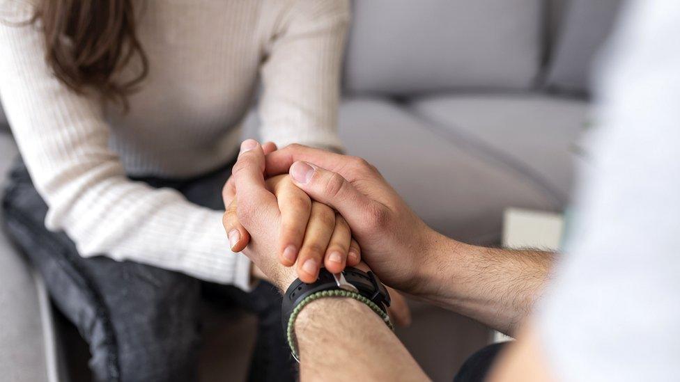 A woman sat on a sofa holding hands with a therapist