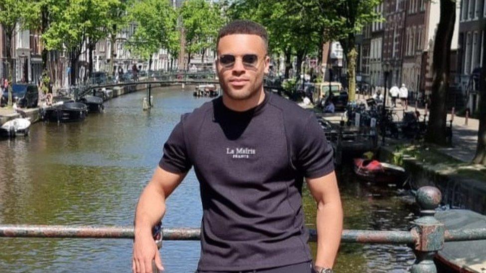 A man wearing sunglasses and a black t-shirt stands on a bridge facing the camera