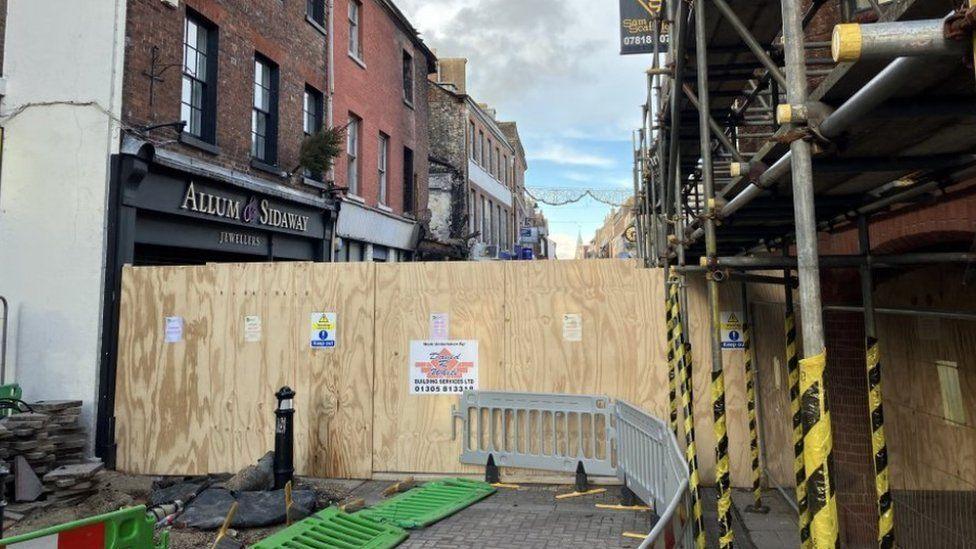 Paved street with buildings either side - scaffolding covers those on the right access is blocked by boarding and plastic fencing.