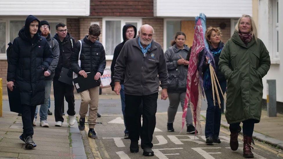 A group of adults in winter clothes walking along a street in Harwich. They are led by a man in a grey jacket with white hair and beard. A woman on teh right is holding a number of flags on poles over her shoulder