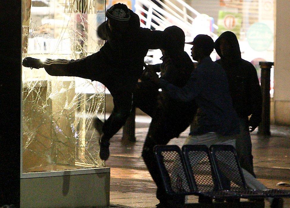 A group of men stand and watch as one man kicks in the window of a jewellers