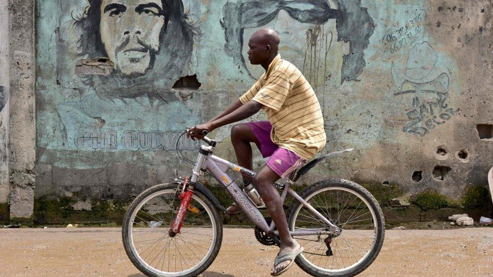 A man on a bike drives past a wall with portraits of Argentine-born guerrilla leader Ernesto "Che" Guevara (1928-1967) (L) and former Libyan dictator Muammar Gaddafi (1942-2011) in a quarter of Abidjan on November 14, 2017.