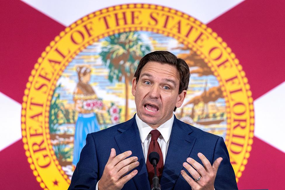 Ron DeSantis stands behind two microphones. He looks impassioned as he gives a speech. He's in front of a picture, surrounded by a golden border with the words "Great Seal of the State of Florida" written in red block capitals