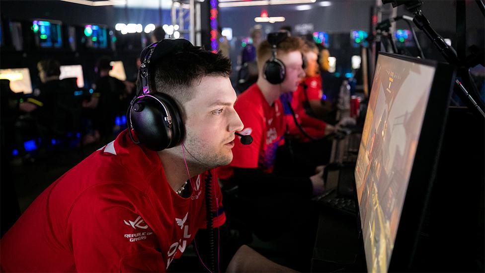 A London Royal Ravens competitor, wearing headphones and a red shirt, playing Call of Duty on a computer screen. There are other team members sitting alongside in the row also playing and wearing red shirts.