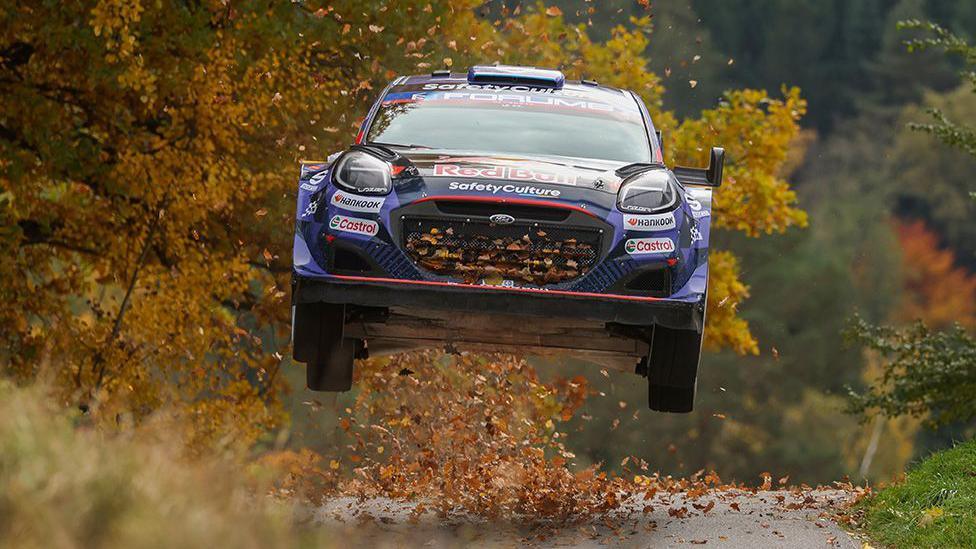 A rally car with Red Bull and sponsor logos is airborne on a narrow road, kicking up autumn leaves. Trees with golden foliage frame the scene, suggesting a forest setting during a race.