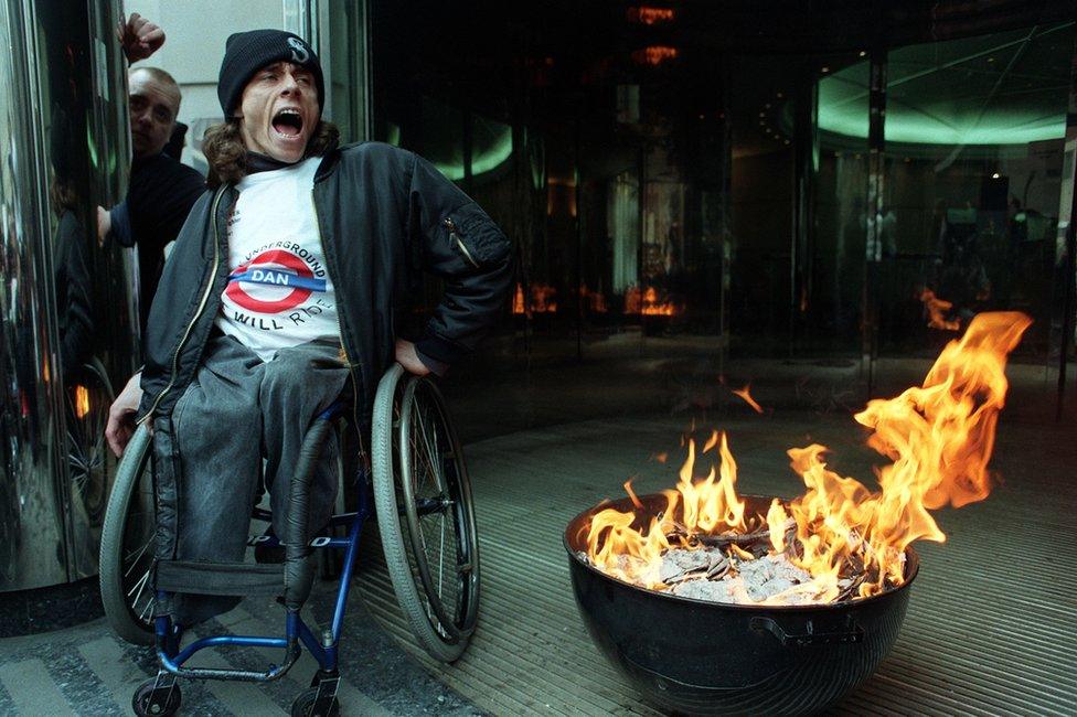 A member of DAN sits beside burning consultation documents, outside the Department of Transport and the Environment in Westminster, central London