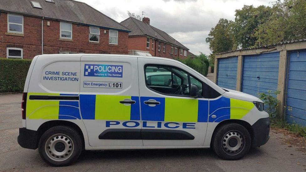 A police van is parked by some blue garages in a car park next to several houses.