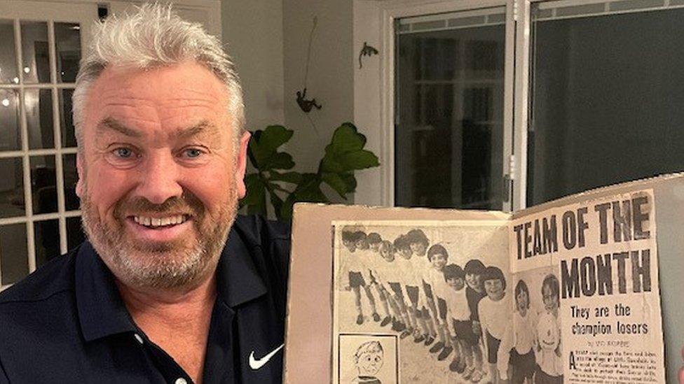 Smiling man with beard holds newspaper cutting showing children's football team