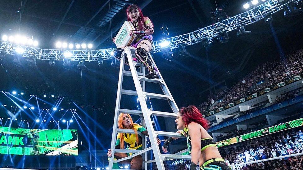 Wrestler Iyo Sky perches on top of a stepladder in the middle of the ring during the women's Money in the Bank match. She's clutching a briefcase, with Money in the Bank written on it. Becky Lynch is handcuffed to the bottom of the ladder at the left while Bayley is handcuffed to the opposite side.