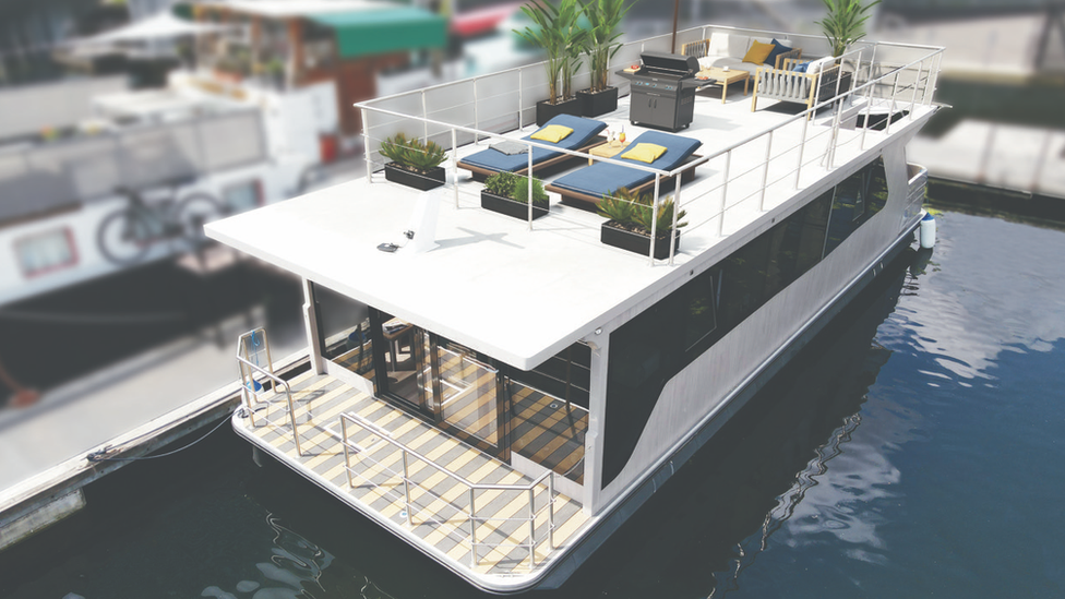 Exterior of boat-like floating home with 2 storeys including a sundeck on top with sunloungers and plants next to a balcony