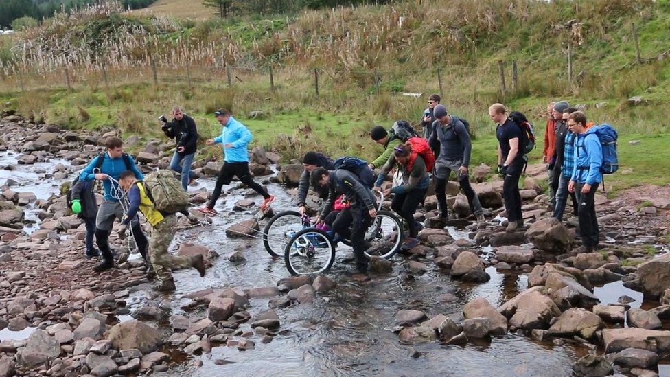 Supporters help Imogen to the top of Pen Y Fan by carrying her across a stream in her bike