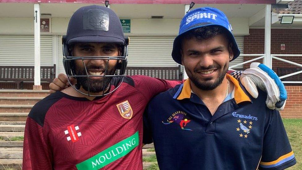 Mohammed on the left and Yash on the right on the pitch at Slough Cricket Club posing for the camera with their arms around each other's shoulder. Mohammed is wearing a navy blue cricket helmet and has a dark beard and has a red cricketing top on as well as a white batting glove on his left hand. Yash has a blue bucket hat on that says "London spirit" on it and is wearing a navy blue polo top. Behind them you can see the front of the cricket pavilion which has some brick steps up to it and some brown benches in front of white shutters.