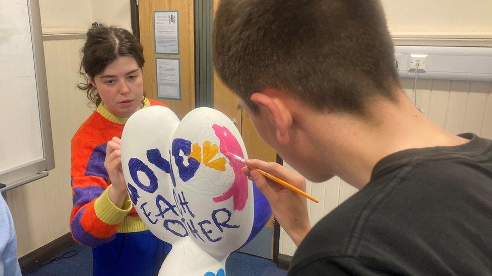 A man and a woman paint the message "love each other" onto a balloon dog sculpture