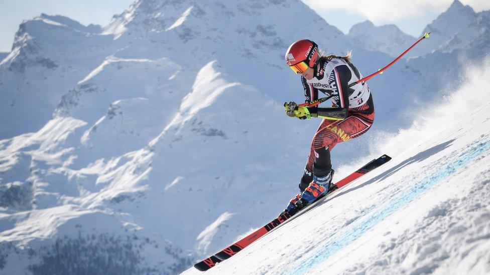 A female downhill skier is seen partially suspended in the air mid-race against the backdrop of snowy mountains