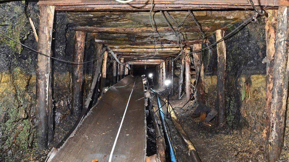 Inside the Gleision drift mine, with wires and a conveyor belt inside a rock walls held up by wooden beams going deep underground.