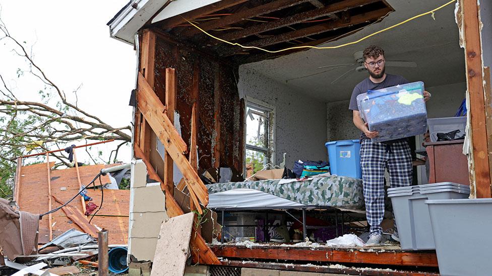 A person collects their things from a bedroom, which is missing a wall due to tornado damage