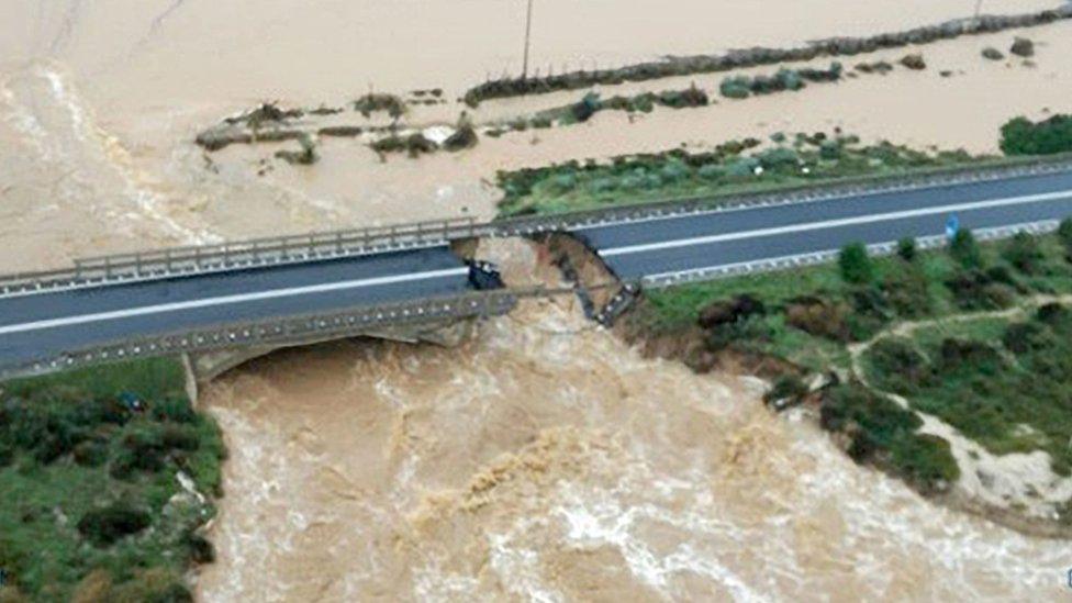 An aerial shot shows a bridge, collapsed in its centre with raging brown water below