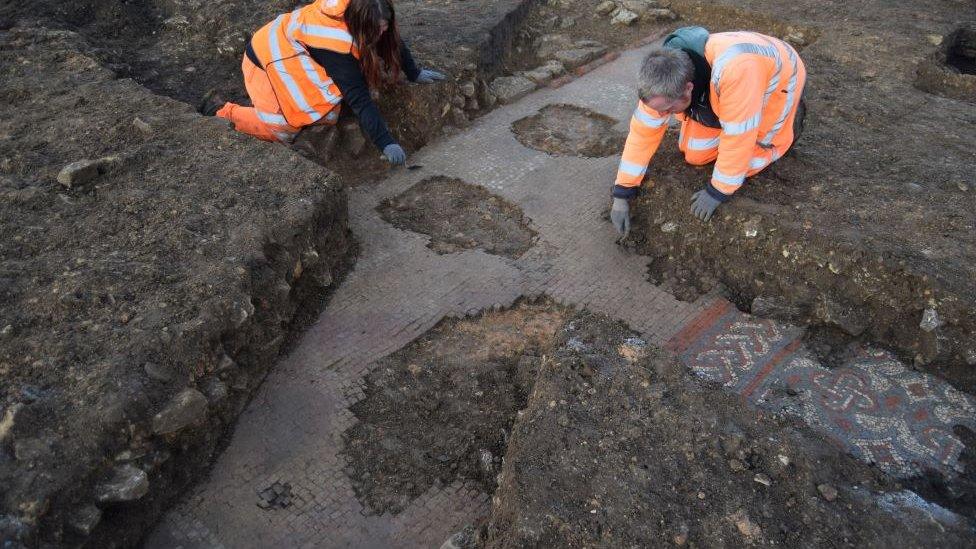 Two archaeologist in high viz outfits carefully excavate the mosaic.