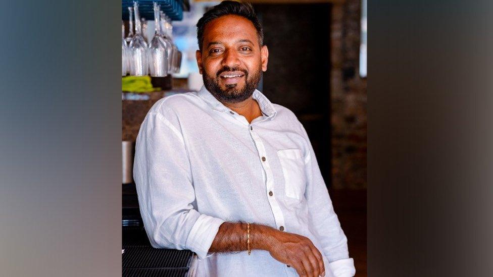 Raja Munuswamy stood leaning on a bar smiling for the camera. He is an Asian middle-aged man and has short dark hair and a dark beard. He is wearing a white shirt with the right sleeve rolled up to his elbow. He has a gold bracelet on his right arm and you can see some wine glasses hanging up above the bar behind him.