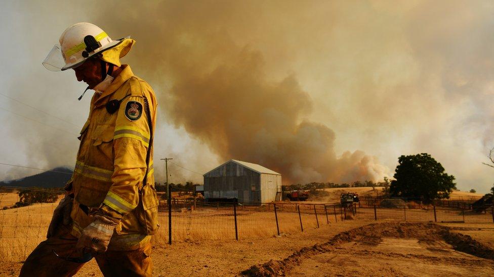 A firefighter in the foreground of the photo with a plume of smoke rising above a barn in the background